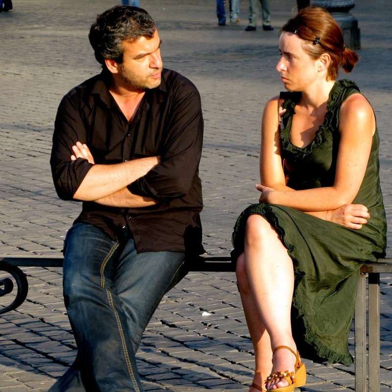 A couple sit next to each other on a public bench with arms and legs crossed in a defensive posture. They stare at each other with frowning faces. A couple sit next to each other on a public bench with arms and legs crossed in a defensive posture. They stare at each other with frowning faces.