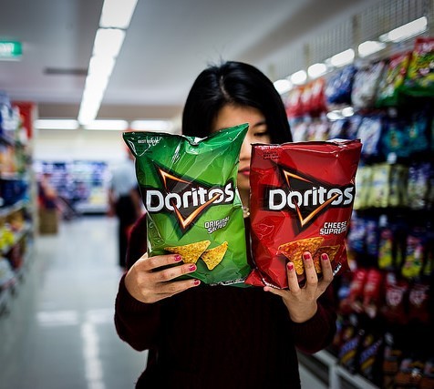 A woman in a supermarket aisle holds two different types of Doritos and carefully reads the packages. A woman in a supermarket aisle holds two different types of Doritos and carefully reads the packages.