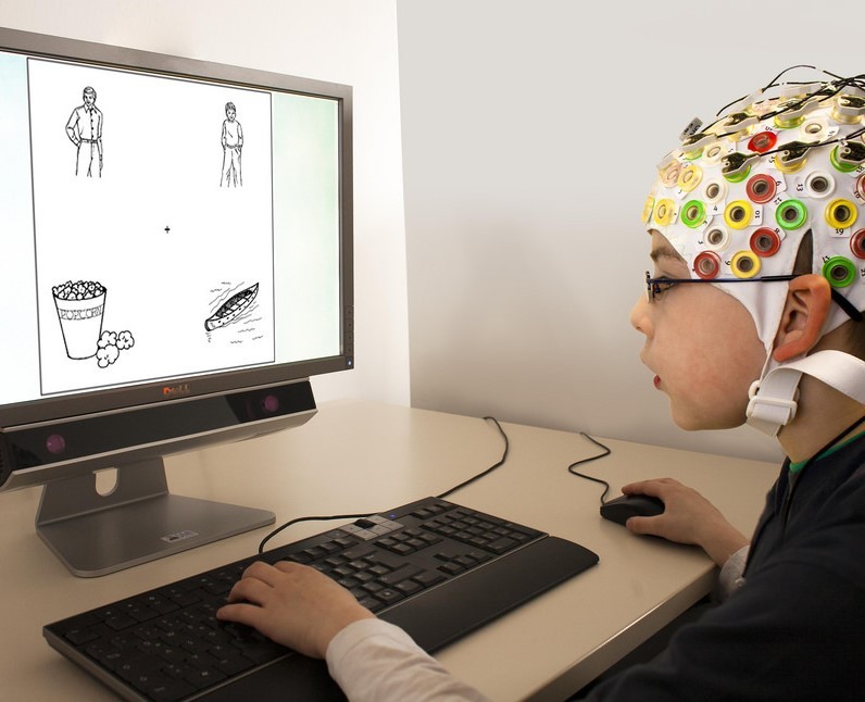 A boy wearing an EEG cap uses a mouse and keyboard while looking at images on a computer monitor during an experiment. A boy wearing an EEG cap uses a mouse and keyboard while looking at images on a computer monitor during an experiment.