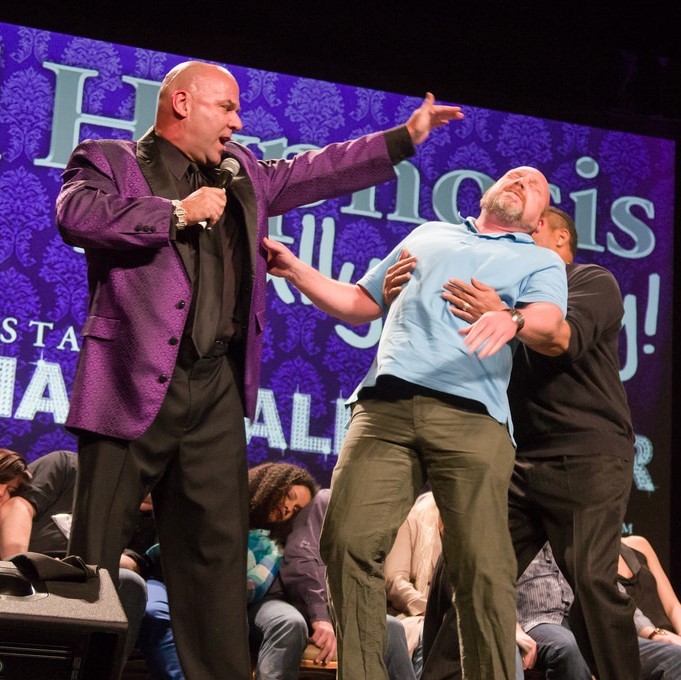 A stage hypnotist holds his hand over the head of a volunteer who falls limp into the arms of the hypnotist's assistant. A group of volunteers seem to be unconscious in their seats in the background. A stage hypnotist holds his hand over the head of a volunteer who falls limp into the arms of the hypnotist's assistant. A group of volunteers seem to be unconscious in their seats in the background.