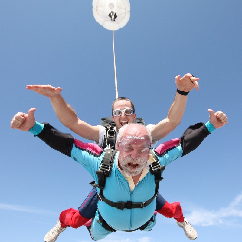 An older man with white hair and beard skydives in tandem with a younger jumpmaster. An older man with white hair and beard skydives in tandem with a younger jumpmaster.