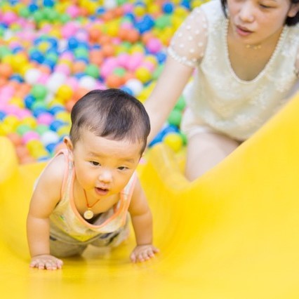 A toddler crawls up a slide in a play area as its mother stands close behind. A toddler crawls up a slide in a play area as its mother stands close behind.