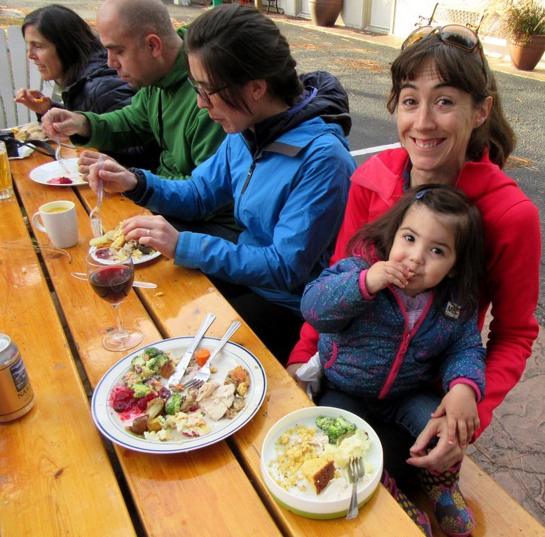 A young girl sits in her mother's lap during a holiday meal as a group of adults around the table enjoy food and drinks. A young girl sits in her mother's lap during a holiday meal as a group of adults around the table enjoy food and drinks.