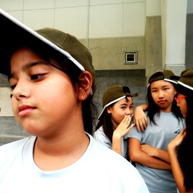 A sad looking girl stands in the foreground as a group of her classmates stands behind looking at her and whispering behind their hands. A sad looking girl stands in the foreground as a group of her classmates stands behind looking at her and whispering behind their hands.