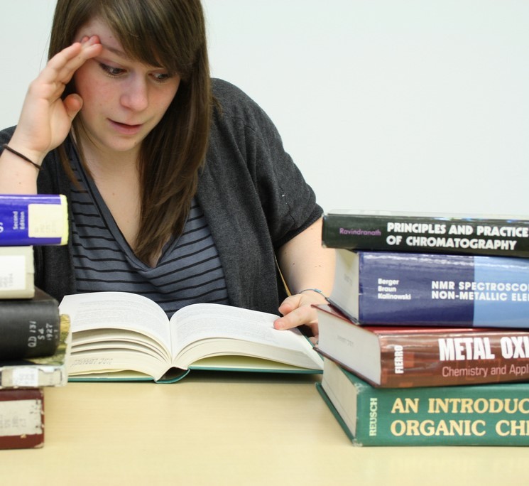 A student reads a large textbook with stacks of other textbooks on the table around her. A student reads a large textbook with stacks of other textbooks on the table around her.