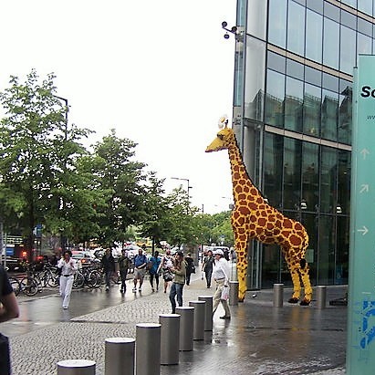 A life-sized model of a giraffe stands in a busy public plaza. A life-sized model of a giraffe stands in a busy public plaza.