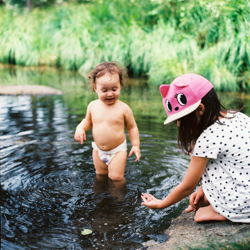 A toddler in a diaper wades in a pond while his sister picks up pebbles on the bank. A toddler in a diaper wades in a pond while his sister picks up pebbles on the bank.