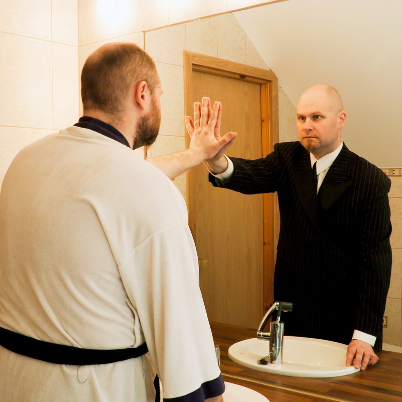 A man stands in front of the bathroom mirror and reaches out to touch an altered reflection of himself. A man stands in front of the bathroom mirror and reaches out to touch an altered reflection of himself.