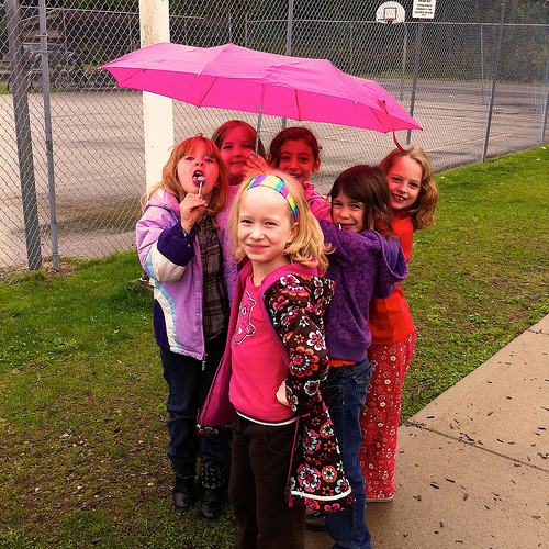 A group of young girls stand together under an umbrella on the playground. A group of young girls stand together under an umbrella on the playground.