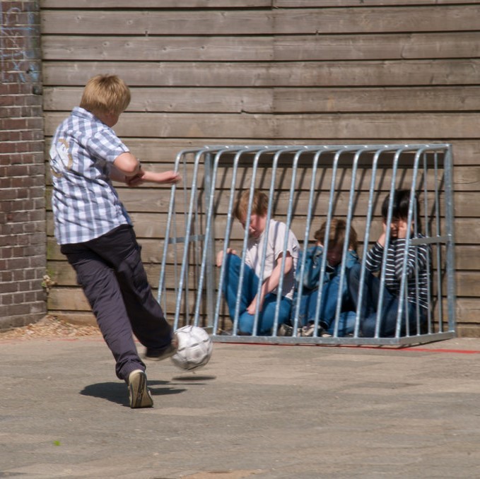 A playground bully kicks a soccer ball at a group of students huddled behind a barrier. A playground bully kicks a soccer ball at a group of students huddled behind a barrier.