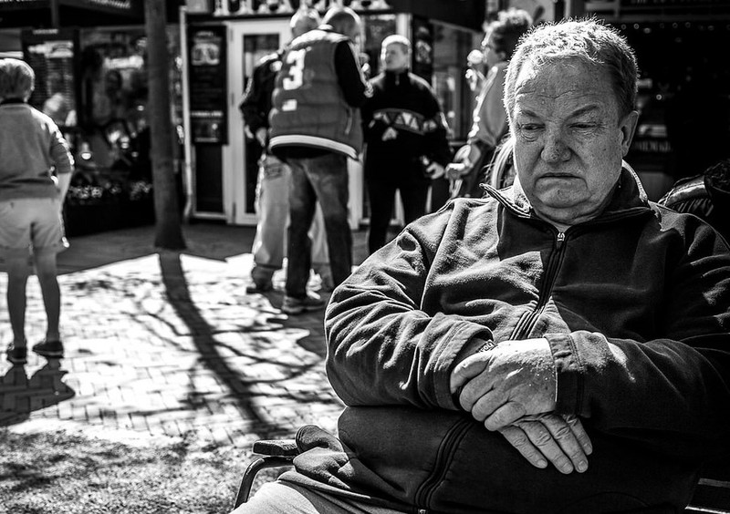 A man sits alone on public bench with his arms crossed and downcast eyes. A man sits alone on public bench with his arms crossed and downcast eyes.