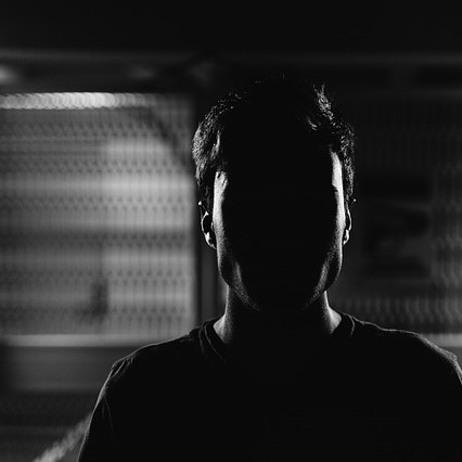 A man stands in a darkened room with his face almost entirely obscured by shadow. A man stands in a darkened room with his face almost entirely obscured by shadow.