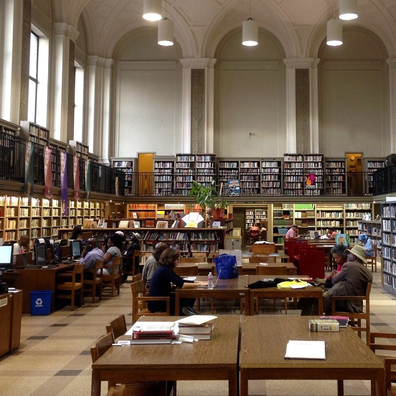 Tall cases of books surround a central area full of desks as people sit reading and accessing information from computers in a public library. Tall cases of books surround a central area full of desks as people sit reading and accessing information from computers in a public library.