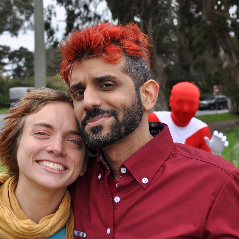A couple pose for a picture in the foreground as a man in a red bodysuit and mask photo bombs them in the distance. A couple pose for a picture in the foreground as a man in a red bodysuit and mask photo bombs them in the distance.