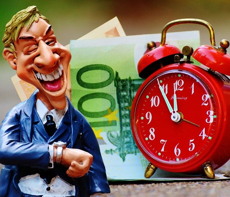 A business man stands grinning and looking at his watch with a clock and some cash visible in the background. A business man stands grinning and looking at his watch with a clock and some cash visible in the background.