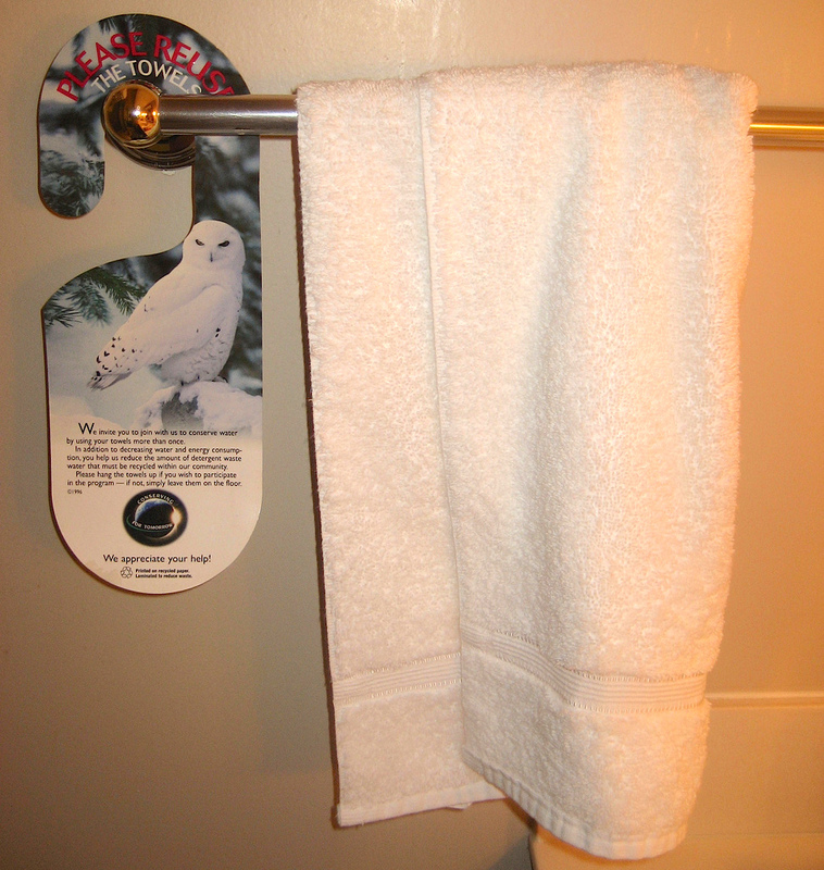 A towel rack in a hotel guest bathroom has a white towel hanging next to an informational sign about how to save water. A towel rack in a hotel guest bathroom has a white towel hanging next to an informational sign about how to save water.