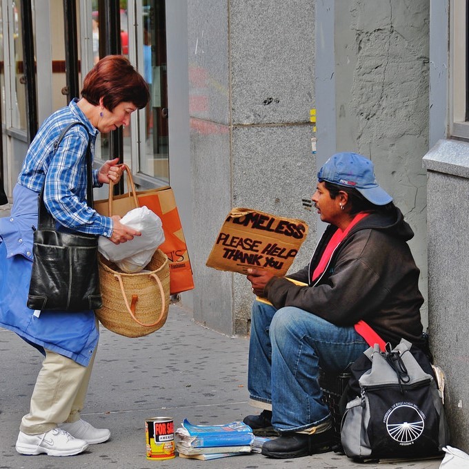 A woman stops on the sidewalk to offer food to a man holding a sign reading "Homeless, please help Thank you." A woman stops on the sidewalk to offer food to a man holding a sign reading "Homeless, please help Thank you."