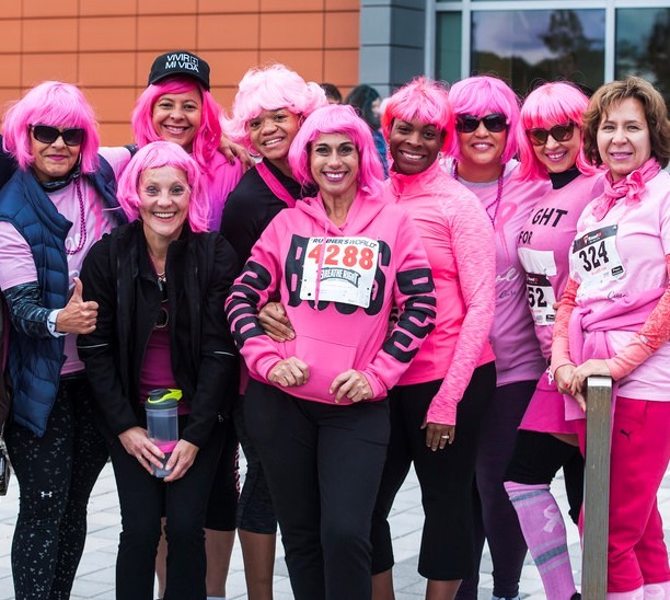 A group of women wearing pink wigs and pink shirts pose together at the conclusion of a 5K race in support of those with breast cancer. A group of women wearing pink wigs and pink shirts pose together at the conclusion of a 5K race in support of those with breast cancer.