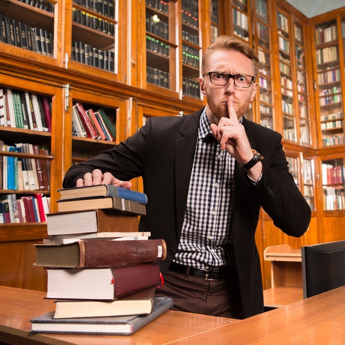 A man dressed in a jacket and bow tie stands behind a desk stacked with books. He has a stern look on his face and is putting a finger to his lips requesting quiet. Behind the man are shelves lined with books. A man dressed in a jacket and bow tie stands behind a desk stacked with books. He has a stern look on his face and is putting a finger to his lips requesting quiet. Behind the man are shelves lined with books.