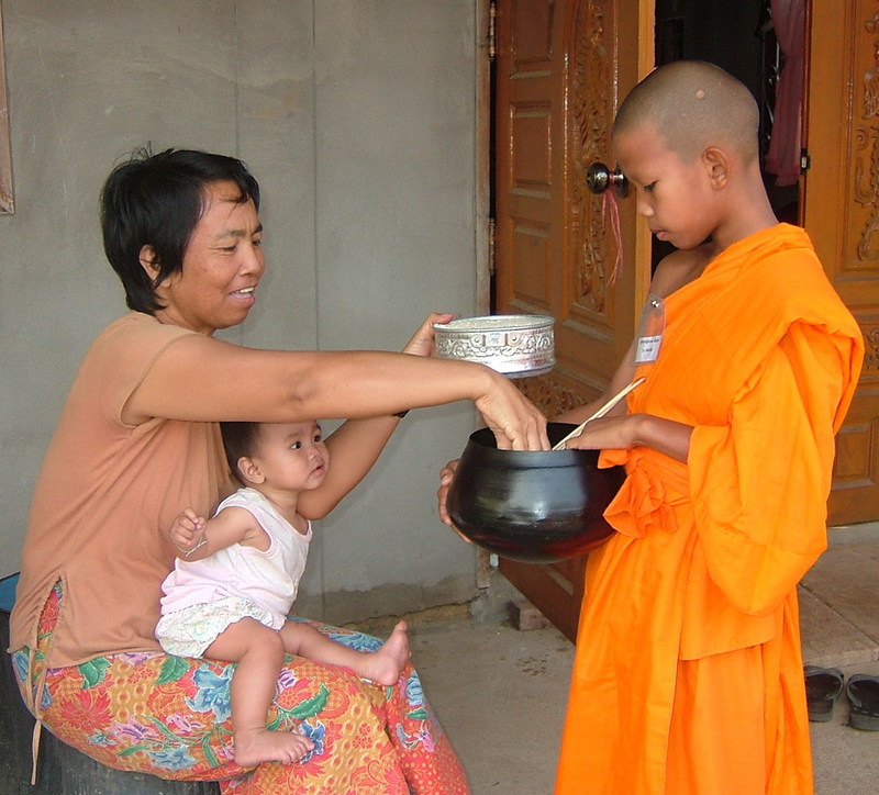 A Buddhist woman with a baby on her lap places food into the alms bowl of a young Buddhist priest dressed in traditional orange robes. A Buddhist woman with a baby on her lap places food into the alms bowl of a young Buddhist priest dressed in traditional orange robes.