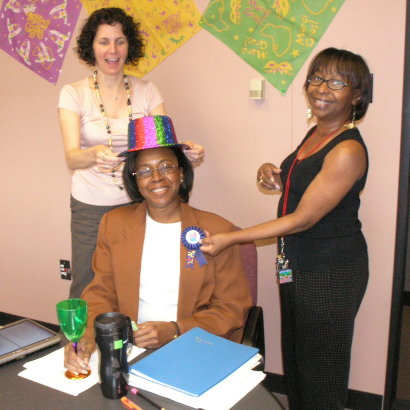 A pair of coworkers put a colorful party hat and ribbon on a colleague as part of an office birthday celebration. A pair of coworkers put a colorful party hat and ribbon on a colleague as part of an office birthday celebration.