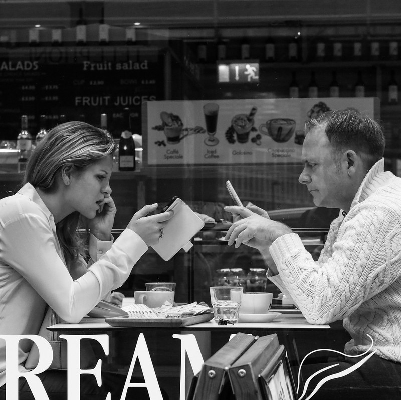 A man and a woman sit across from one another at a small table in a coffee shop. Both of them are staring at their own smartphone rather than engaging with each other. A man and a woman sit across from one another at a small table in a coffee shop. Both of them are staring at their own smartphone rather than engaging with each other.