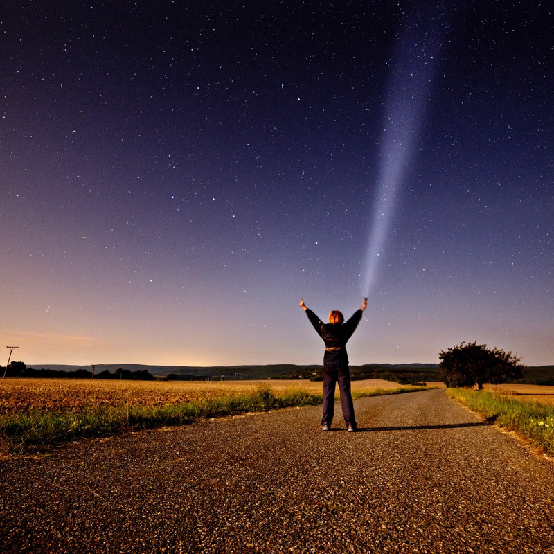 A woman stands in the middle of a country road at night and reaches toward the star-filled sky above. A woman stands in the middle of a country road at night and reaches toward the star-filled sky above.