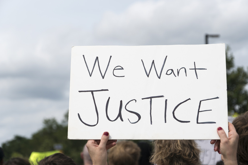 This image depicts two hands holding a sign high. The sign is white and has a hand-written message that reads "We want justice" in black letters. In the distance, one can see the fuzzy images of many heads, suggesting that this sign is being held up at a gathering such as a protest. There are trees in the distance. This image depicts two hands holding a sign high. The sign is white and has a hand-written message that reads "We want justice" in black letters. In the distance, one can see the fuzzy images of many heads, suggesting that this sign is being held up at a gathering such as a protest. There are trees in the distance.