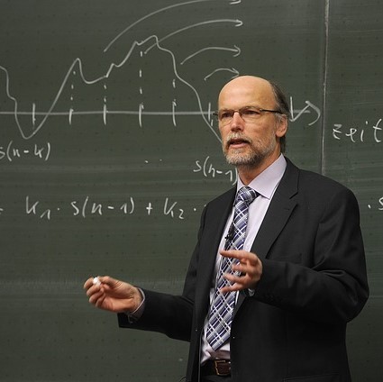 A stereotypical image of a professor - a white, middle-aged man with glasses and a beard, dressed in a coat and tie stands with chalk in hand in front of a blackboard which displays a mathematical formula. A stereotypical image of a professor - a white, middle-aged man with glasses and a beard, dressed in a coat and tie stands with chalk in hand in front of a blackboard which displays a mathematical formula.