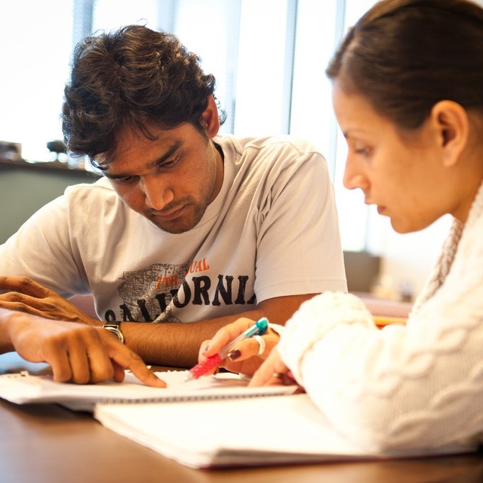 A male and female student work together at a table and focus on details in a notebook in front of them. A male and female student work together at a table and focus on details in a notebook in front of them.