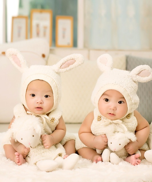 Twin boys sit together dressed in matching clothes and hats and holding similar stuffed animals. Twin boys sit together dressed in matching clothes and hats and holding similar stuffed animals.