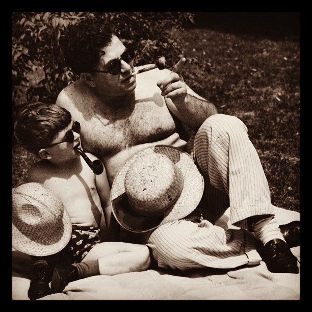 A father and his young son sit together on a blanket on the lawn on a sunny day. Each have their shirts removed and are dressed almost identically including straw hats, sunglasses, and pipes. A father and his young son sit together on a blanket on the lawn on a sunny day. Each have their shirts removed and are dressed almost identically including straw hats, sunglasses, and pipes.