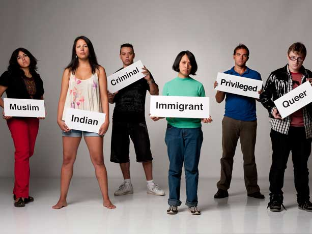 A group of people stand holding signs labeling them as others perceive them. The signs include, "Muslim", "Indian", "Criminal", "Immigrant", "Privileged", and "Queer". A group of people stand holding signs labeling them as others perceive them. The signs include, "Muslim", "Indian", "Criminal", "Immigrant", "Privileged", and "Queer".