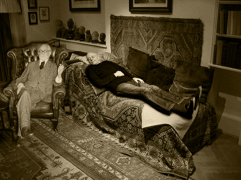 This is a black and white image of Sigmund Freud conducting psychoanalysis with a patient. The image shows Freud, wearing a suit and glasses, sitting in a leather chair facing away from the patient. The patient, a middle-aged man, is lying on a couch that is covered by a patterned blanket that is similar to a number of oriental rugs covering the floor. A number of framed photographs and books can be seen in the background. This is a black and white image of Sigmund Freud conducting psychoanalysis with a patient. The image shows Freud, wearing a suit and glasses, sitting in a leather chair facing away from the patient. The patient, a middle-aged man, is lying on a couch that is covered by a patterned blanket that is similar to a number of oriental rugs covering the floor. A number of framed photographs and books can be seen in the background.